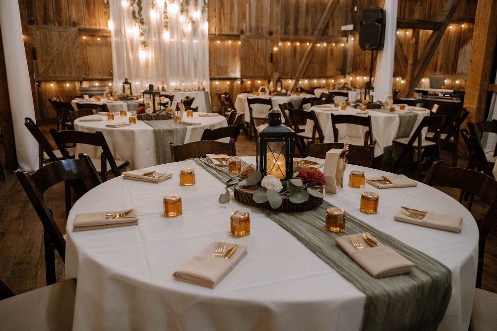 Elegant barn wedding reception tables set with white linens, lanterns, and string lights overhead.