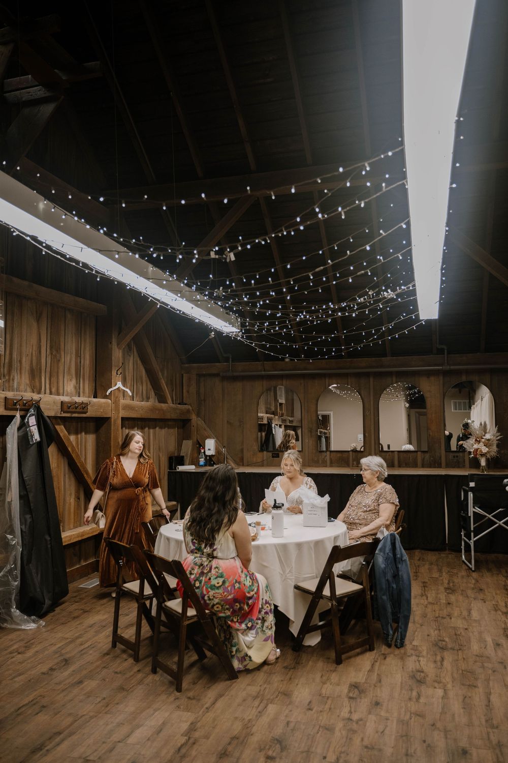 Women gathered at a round table inside a rustic barn venue decorated with fairy string lights.
