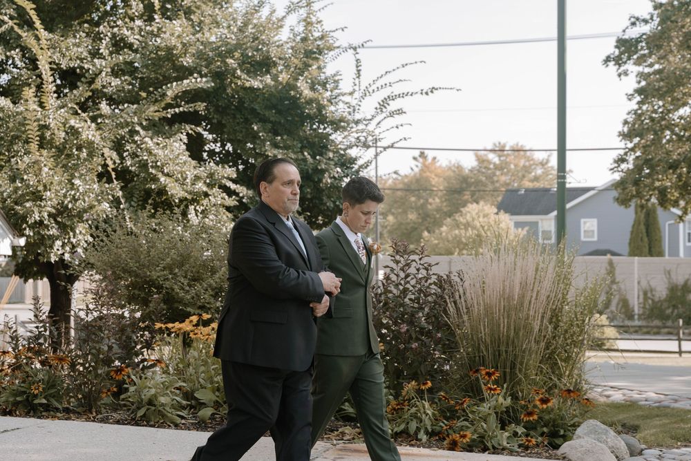 Two men in suits walk together along a garden path at a wedding event.