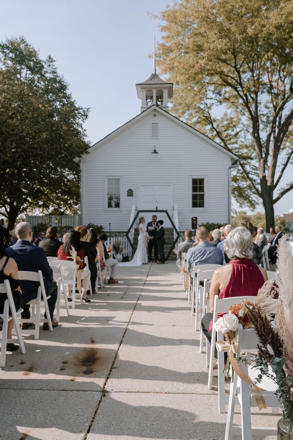Outdoor wedding ceremony at a white country church with guests seated in white chairs along an aisle on a sunny day.