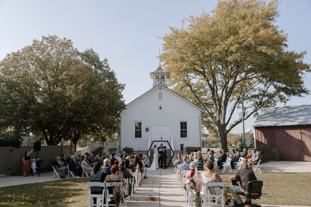 Wide-angle view of an outdoor wedding ceremony at a charming white chapel surrounded by autumn trees and seated guests.