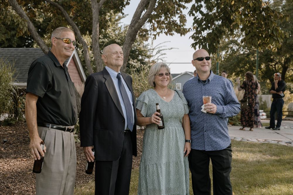 Four adults posing outdoors at a casual garden gathering, dressed in semi-formal attire near trees.