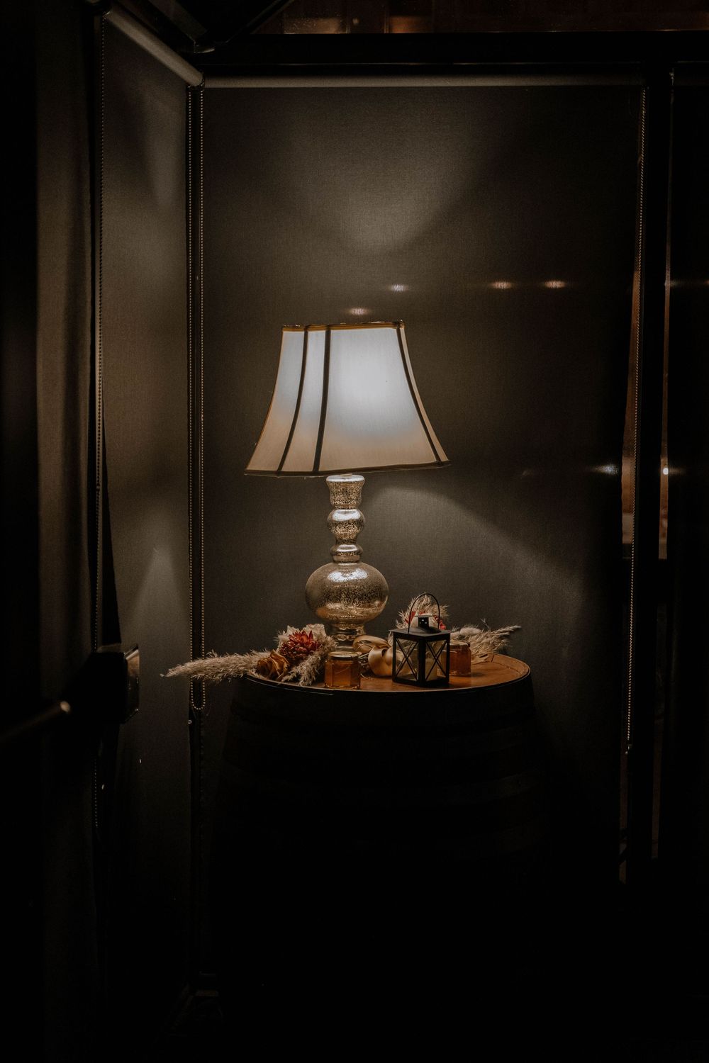 A glowing table lamp with a white shade on a wooden side table in a dark, moody room with decorative accents.