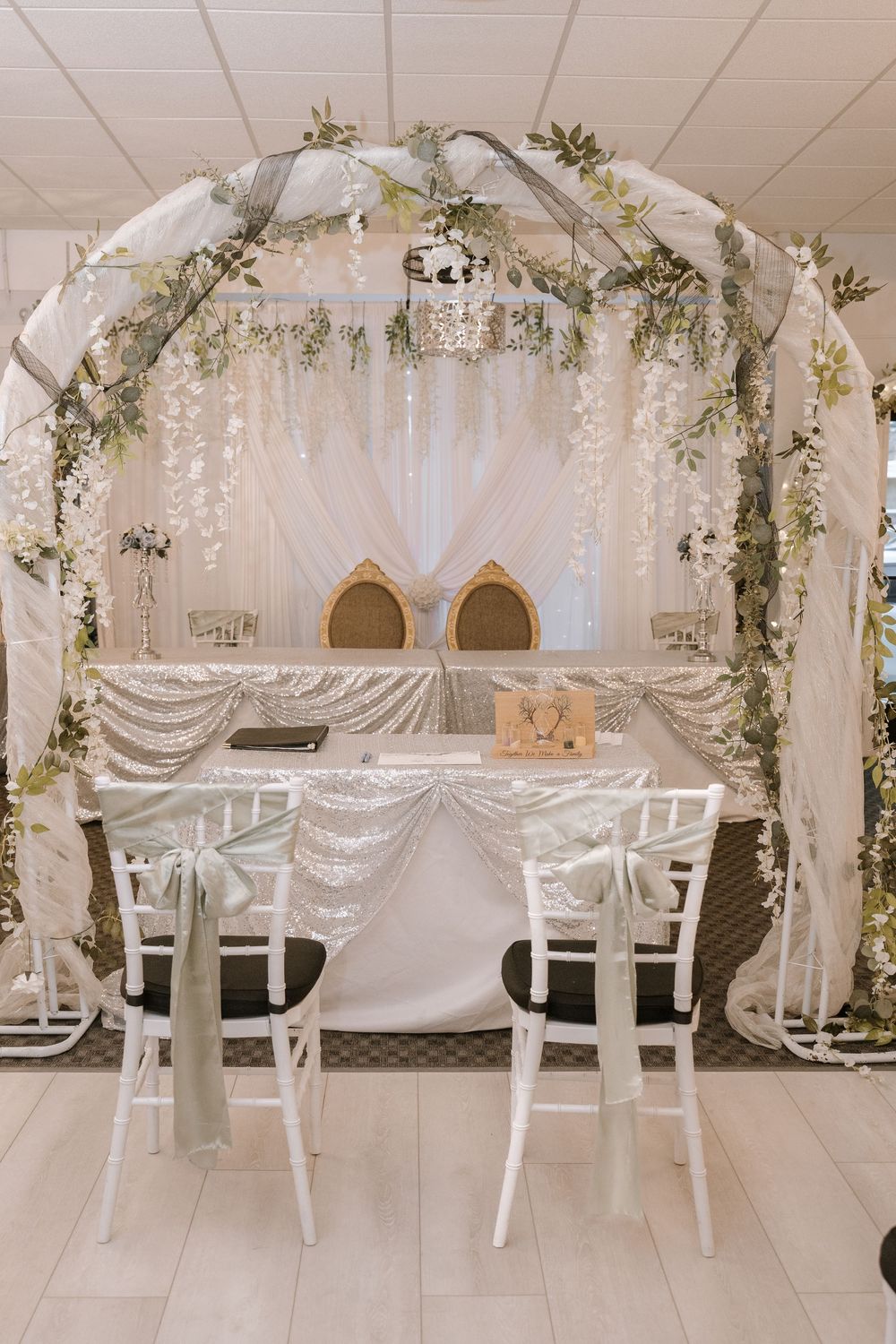 Elegant wedding chuppah with white draping, greenery, floral accents, two chairs, and a decorated table for a Jewish ceremony.