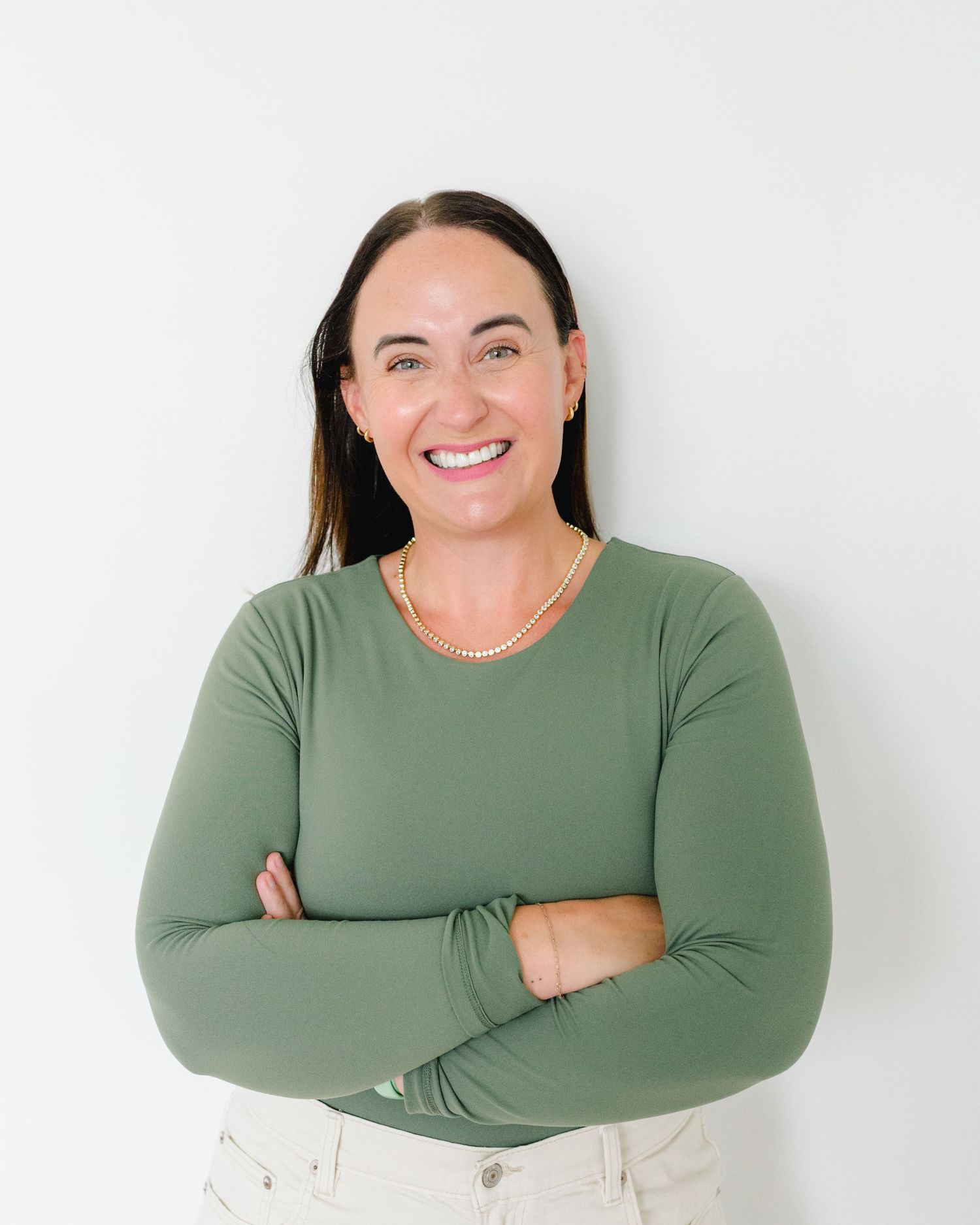 Smiling woman in green long-sleeve top with arms crossed, wearing a gold necklace, posed against a white background.