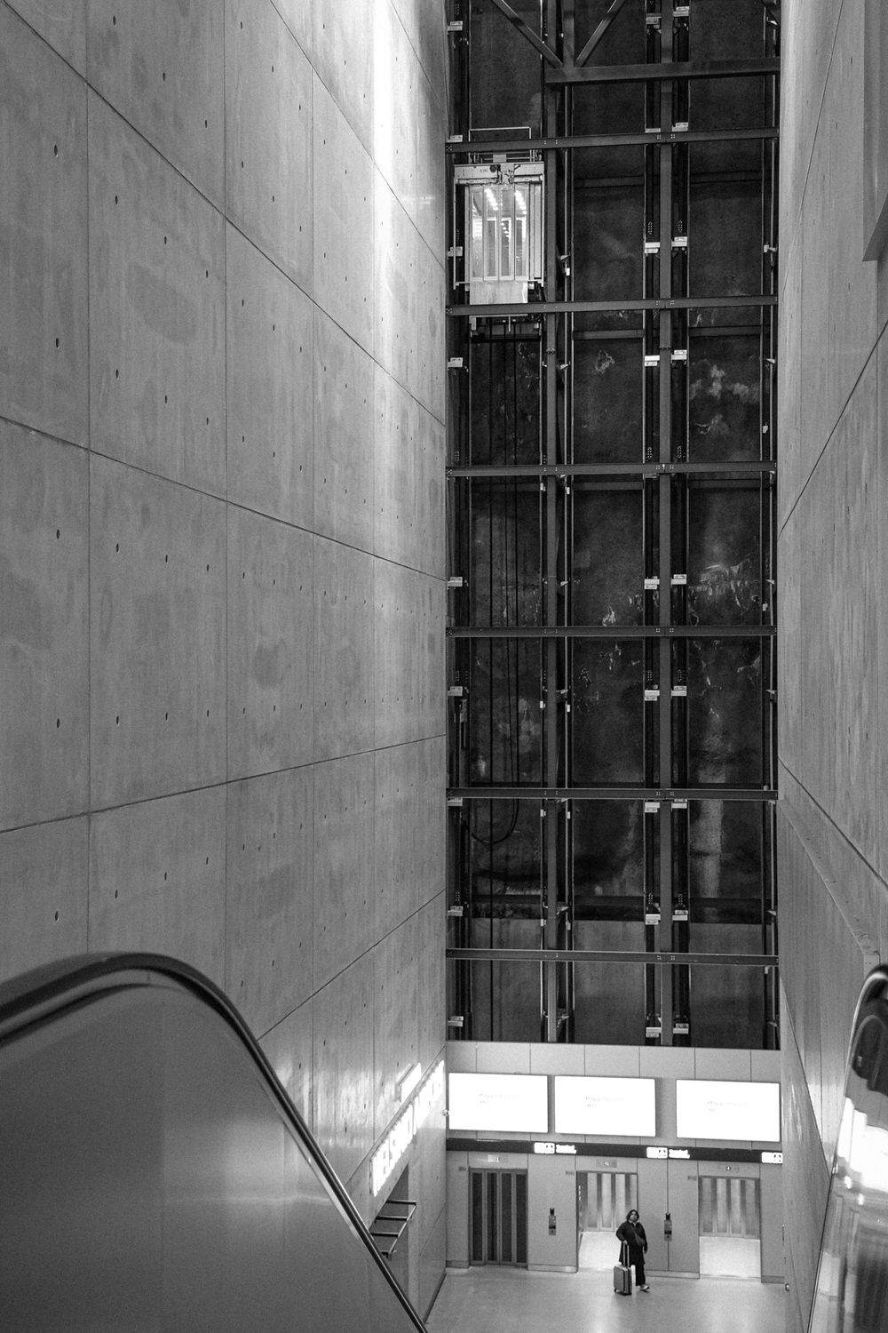 Black and white photo of a modern building interior with escalator, concrete walls, and tall glass facade.