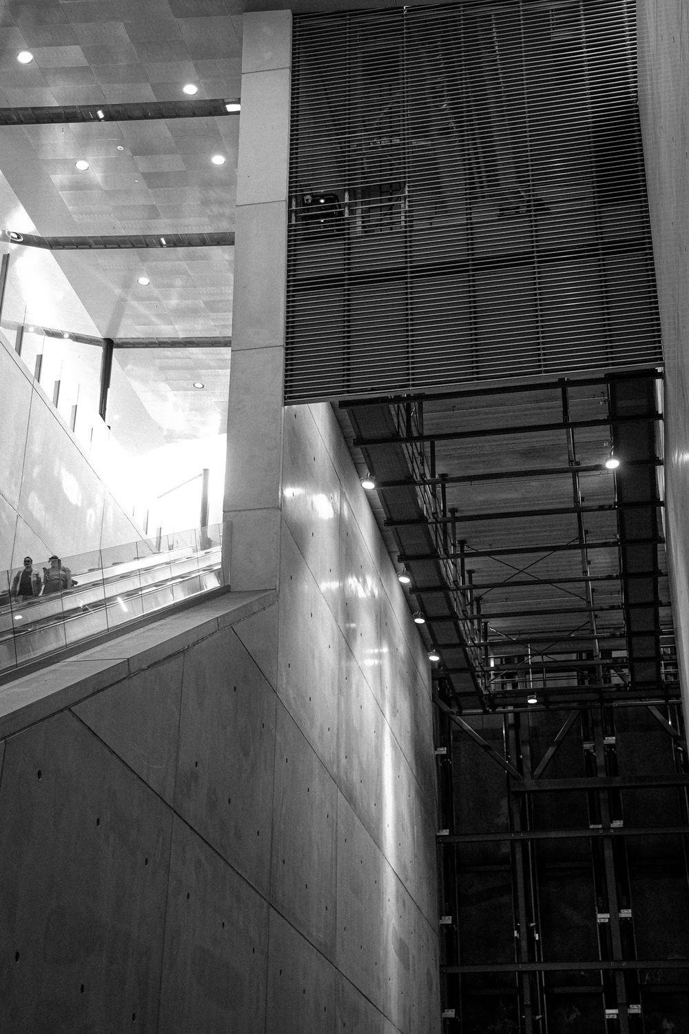 Black and white architectural interior featuring concrete walls, glass panels, escalators, and dramatic lighting in a modern building.
