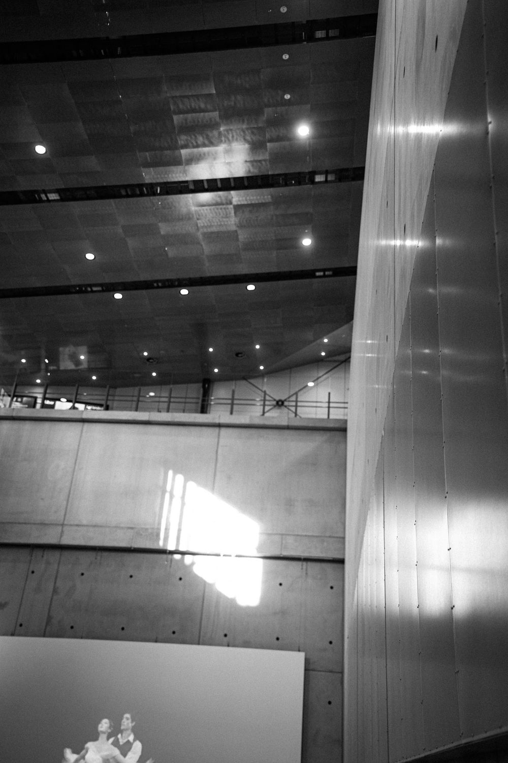 Black and white photo of a Helsinki Airport railway station concourse with reflective floors, columns, and a Railway Station sign.