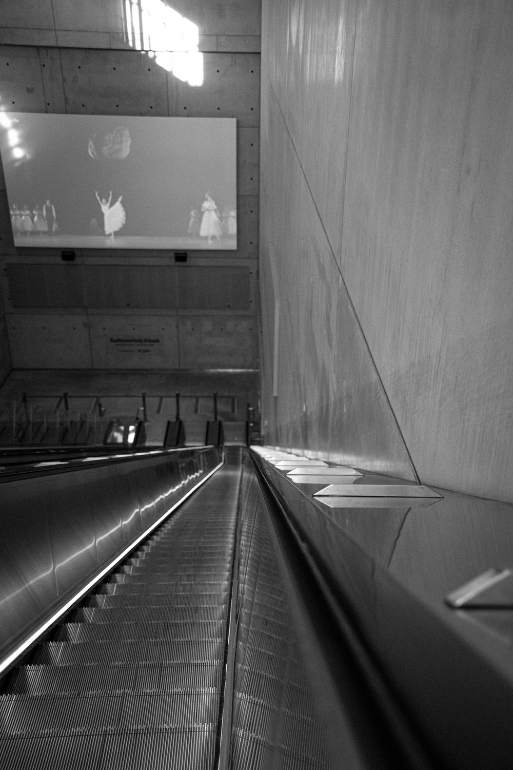 Black and white photo of a Helsinki Airport railway station concourse with reflective floors, columns, and a Railway Station sign.