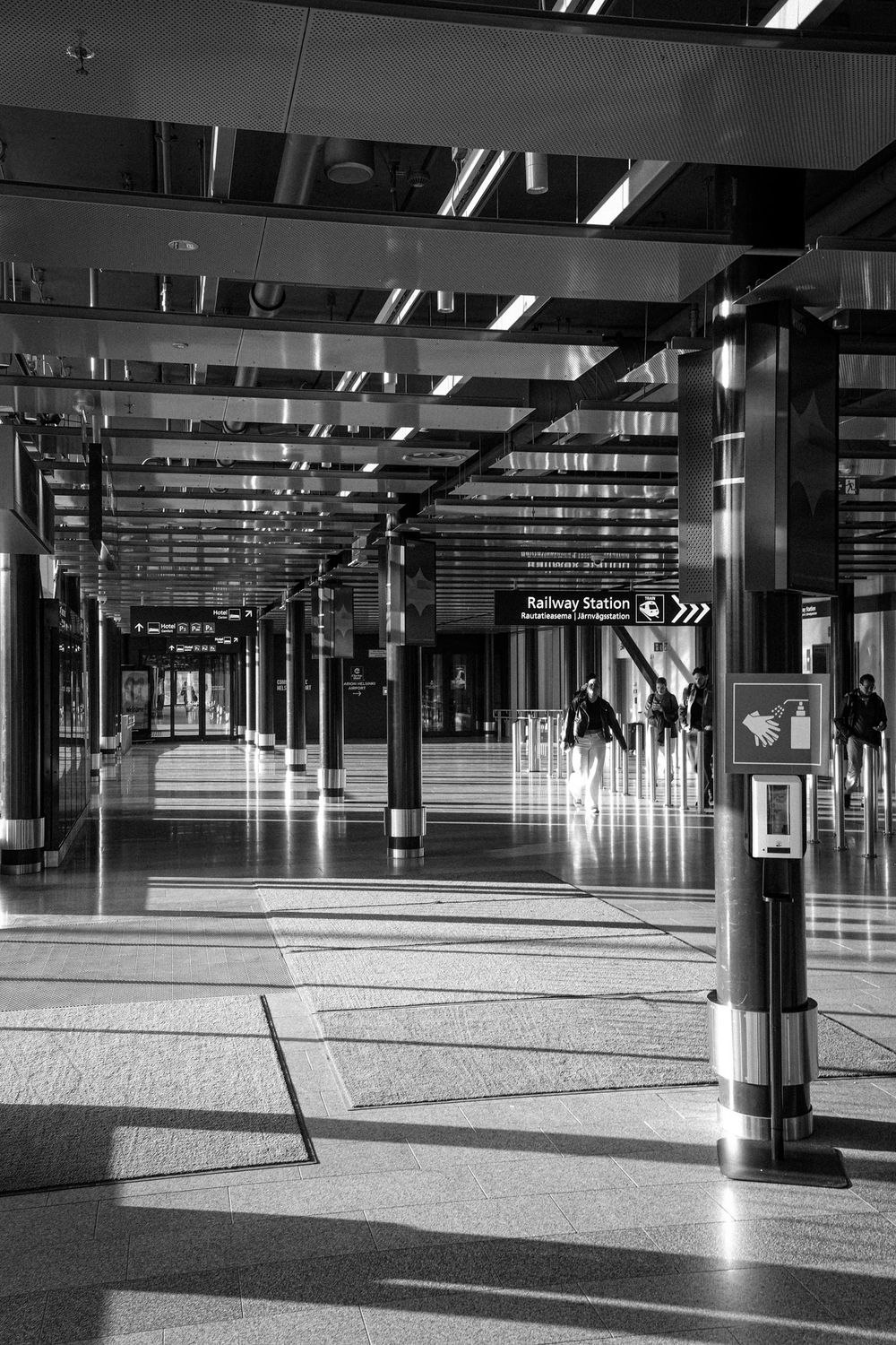 Black and white photo of a Helsinki Airport railway station concourse with reflective floors, columns, and a Railway Station sign.