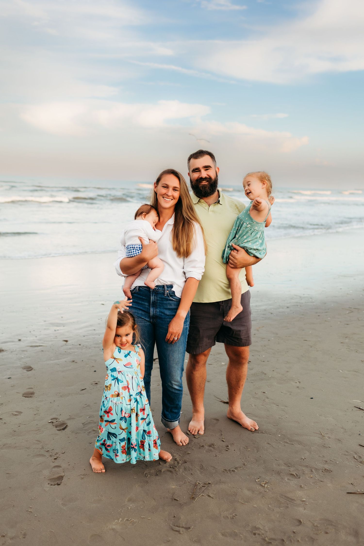 Family of five posing on a sandy beach at sunset, parents holding toddlers with young girl standing in front.