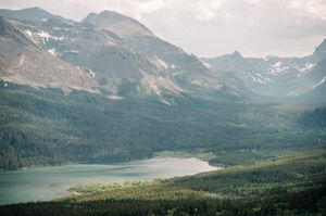 Aerial view of a serene mountain lake surrounded by dense evergreen forest and snow-capped peaks in a scenic valley.