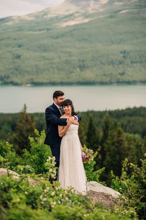 Bride and groom embrace on a lush hillside with a serene mountain lake and forest backdrop.
