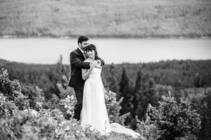 Black and white wedding photo of couple embracing on rocky overlook with forest and lake in background.
