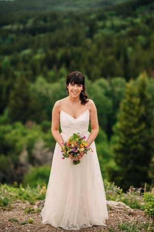Bride in white spaghetti strap wedding gown holding colorful bouquet with mountain forest backdrop.