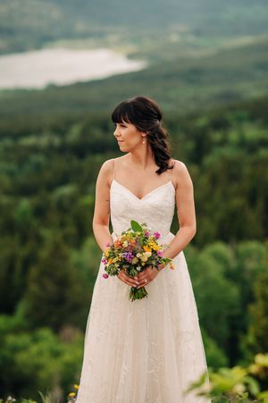 Bride in white lace gown holds colorful bouquet with mountain forest and lake backdrop.