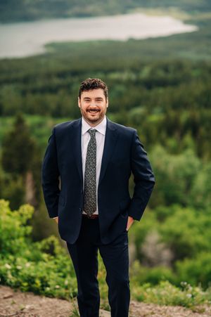 Smiling groom in navy suit stands confidently with scenic mountain lake background.