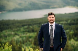 Professional man in navy suit and tie smiling outdoors with scenic mountain lake and forest backdrop.