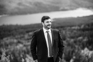 Man in suit smiling outdoors with mountain lake backdrop in black and white.