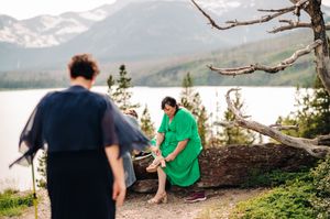 Woman in green dress sits on log by mountain lake as another in navy blue stands in foreground.