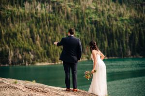 Bride and groom standing at rocky lakeside overlook with lush mountain forest in background.