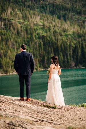 Bride and groom stand at edge of turquoise mountain lake surrounded by evergreen forest during outdoor wedding.