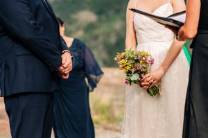 Bride holding a colorful wildflower bouquet stands beside groom in navy suit during an outdoor wedding ceremony.