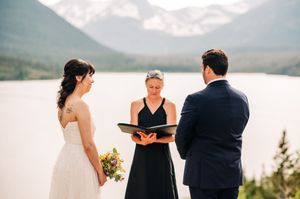 Outdoor wedding ceremony with officiant reading vows beside mountain lake backdrop.