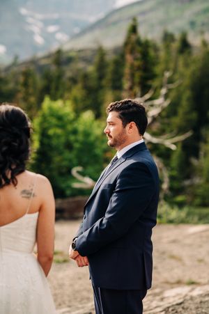Groom in navy suit watches bride during outdoor mountain wedding ceremony with forest backdrop.