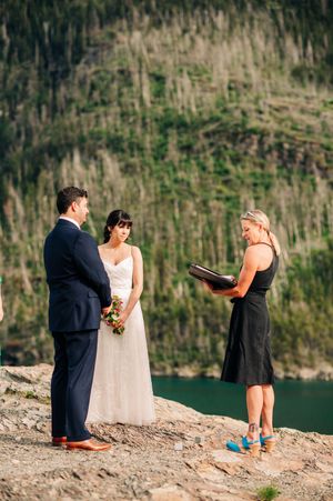Couple exchanges vows with officiant at outdoor mountain lake wedding ceremony on rocky cliff.