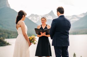 Outdoor wedding ceremony with officiant reading vows before couple with mountain lake backdrop.
