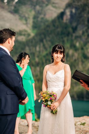 Bride holding colorful wildflower bouquet during outdoor mountain wedding ceremony with turquoise lake backdrop.
