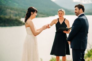 Bride placing wedding ring on groom's finger during lakeside mountain ceremony.