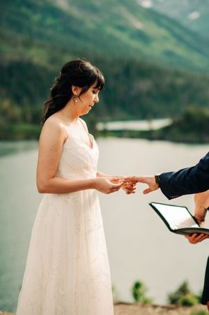 Bride placing wedding ring on groom's finger during outdoor ceremony with mountain lake backdrop.