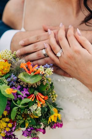 Newlyweds hands showing wedding rings resting on colorful wildflower bridal bouquet.