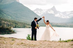 Joyful bride and groom dance together on rocky shoreline with mountain lake scenic backdrop.