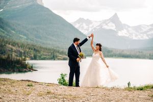 Groom twirling bride in wedding dress by mountain lake with scenic peaks behind them.