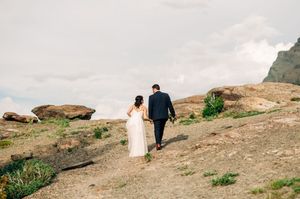 Bride and groom walk hand in hand along rocky mountain trail under cloudy sky.