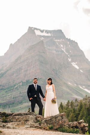 Couple poses on rocky ledge with dramatic snow-capped mountain peak behind them.
