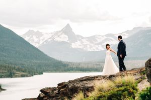 Bride and groom hold hands on rocky cliff overlooking a stunning snow-capped mountain lake.