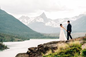 Bride and groom walk hand in hand along rocky cliff with stunning mountain lake landscape.