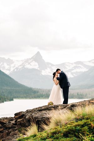 Bride and groom share a romantic kiss on rocky terrain with snow-capped mountains and lake backdrop.