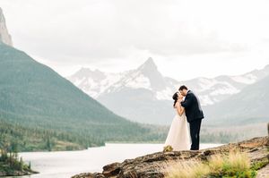 Newlyweds share a kiss on rocky cliff overlooking serene mountain lake and peaks.