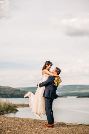 Groom lifts bride in wedding attire by a scenic lake with rolling hills under a cloudy sky.