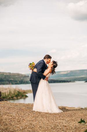 Groom dips bride for a kiss outdoors near a lake, bride holding colorful wildflower bouquet.