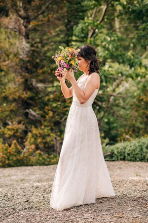 Bride in white lace gown smelling vibrant wildflower bouquet surrounded by autumn forest.