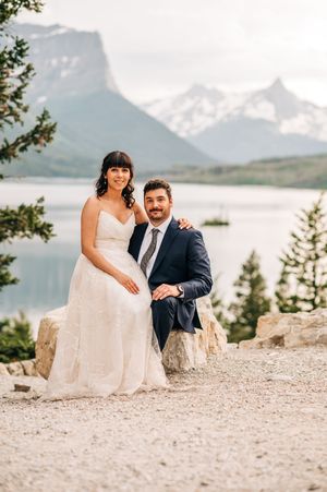 Smiling bride and groom pose together on rocky overlook with mountain lake backdrop at their wedding.