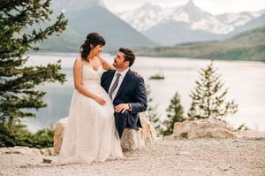 Bride and groom sharing tender moment on rocky overlook with mountain lake and peaks behind.