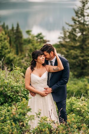 Groom tenderly embraces bride among lush green mountain foliage with misty lake in background.