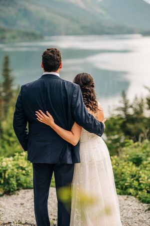 Bride and groom gaze at a serene mountain lake, bride resting hand on grooms back.